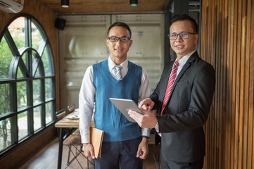 Two Asian businessman standing and looking to bablet, they are meeting and talking about business plan, marketing and financial in the future. they are looking and point finger to detail in notebook.