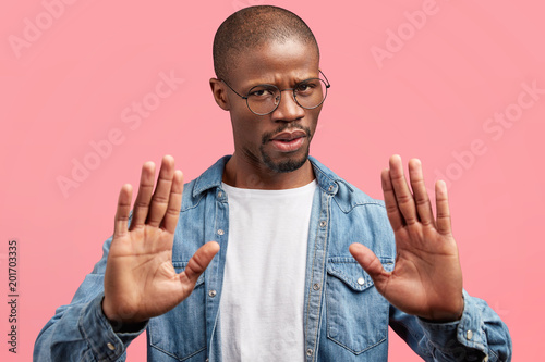 Fotografía Stern dark skinned male boss with serious angry expression, makes stop gesture, tries to protect himself, refuses to sign contract or documents, isolated over pink background