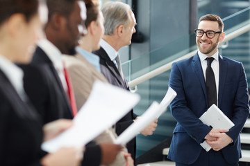 Wall Mural - Successful businessman in elegant formalwear standing in front of business team with papers and listening to their reports at briefing