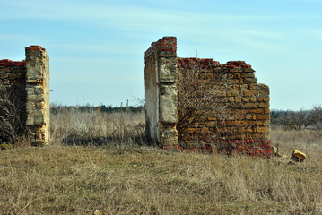 Ruined entrance in farm walls of Crimean coquina rock blocks, dry weathered grass field, blue spring sky background