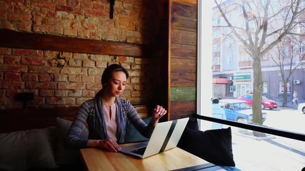 Wall Mural - Pretty female person sitting at cafe on sofa and chatting in social networks by laptop. Concept of typing messages to friends and having leisure time with modern technology. Young woman dressed in