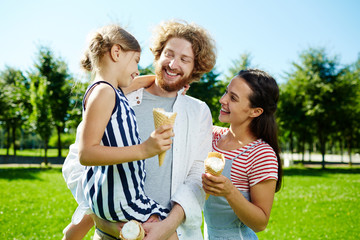 Wall Mural - Happy little girl and her parents with ice creams talking and enjoying sunny day in park