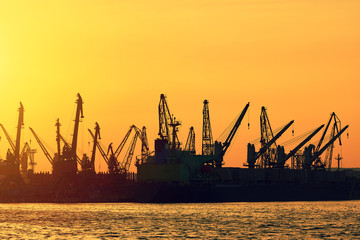 A city landscape. Bright sunset in the seaport. A sunny path on the calm surface of the sea. Large silhouettes of loading cranes. Bright abstract background ideal for any design      