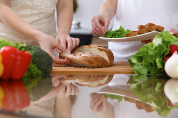 Closeup of human hands cooking in kitchen. Mother and daughter or two female cutting bread at the table full of vegetables and green salad. Healthy meal, vegetarian food and lifestyle concepts