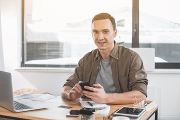 Poster - Portrait of smiling young man typing in mobile while sitting at table in office. Happy employer during job concept