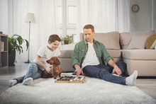 Pensive Man Is Moving Checker On Board And Smiling. His Son Is Sitting Near Him On Carpet And Stroking The Dog
