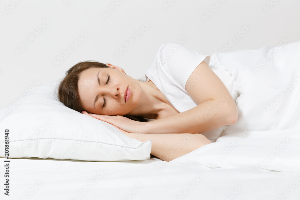 Calm young brunette woman lying in bed with white sheet, pillow, blanket on white background. Sleeping beauty female spending time in room. Rest, relax, good mood concept. Copy space for advertisement - obrazy, fototapety, plakaty 
