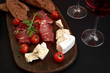 © nazarovsergey - Prosciutto, salami, baguette slices, tomatoes and nutson rustic wooden board, two glasses of red wine over black background