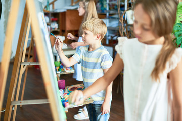side view at group of children painting on easels during art class studio, pretty focus on little bo