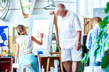 Wall Mural - Portrait of senior art teacher leaning over easel to watch little student painting pictures on easel in art school, copy space