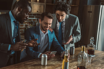 smiling multiethnic male friends in suits using smartphone and drinking alcoholic beverages together