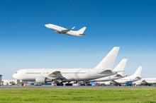 Passenger Aircraft Row, Airplane Parked On Service Before Departure At The Airport, Other Plane Push Back Tow. One Take Off From The Runway In The Blue Sky.