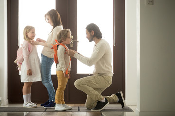 Wall Mural - Caring parents helping happy kids put backpack on preparing go to school standing at house hall together, loving family mom and dad dressing getting children son daughter ready in the morning at home