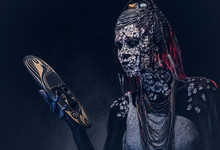 Make-up Concept. Portrait Of A Scary African Shaman Female With A Petrified Cracked Skin And Dreadlocks, Holds A Traditional Mask On A Dark Background. Make-up Concept.