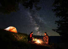 Young Couple Hikers - Male And Female Warming Themselves Around The Campfire At Night Camping Near The Forest And Orange Glowing Tent, Under The Incredible Beautiful Starry Sky And Milky Way.