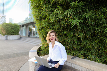 Smiling businesswoman working with laptop and papers in open air near green plant. Concept of busines, modern technology and working online.