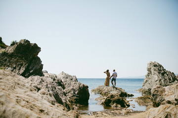 Beautiful romantic couple standing on the rock in the sea with cliffs around. Man and woman holding hands. Greece