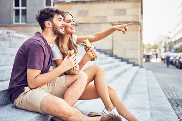 Wall Mural - Young couple eating lunch outdoors sitting on stairs and pointing in the distance