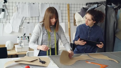 Wall Mural - Young tailors are working with tablet, communicating and outlining clothing pattern on textile on studio desk. Light workshop with sewing items in background.