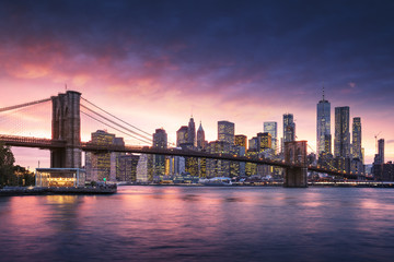 Fototapete - Famous Brooklyn Bridge in New York City with financial district - downtown Manhattan in background. Sightseeing boat on the East River and beautiful sunset over Jane's Carousel.