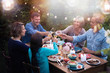 Group of friends in their forties gathered around a table in a garden one summer evening to share a meal. They toast with their glasses of wine