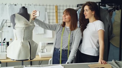 Wall Mural - Two cheerful clothing designers are making funny selfie with smart phone while standing beside clothed mannequin in studio. Attractive women are smiling and posing.