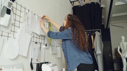 Wall Mural - Low angle view of young fashion designer looking at sketches and hanging drawings on wall in modern loft style studio. Large collection of illustration above tailoring desk.