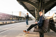 © Maskot - Young man sitting on bench with teenage girl in background at railroad station platform