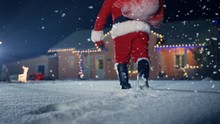 Santa Claus Carrying Red Bag Over The Shoulder, Walks Into Front Yard Of The Idyllic House Decorated With Lights And Garlands. Santa Bringing Gifts And Presents. 