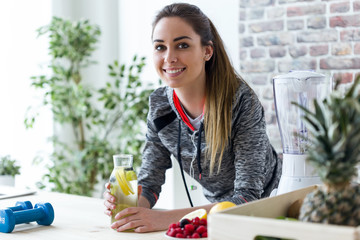 Wall Mural - Sporty young woman looking at camera while drinking lemon juice in the kitchen at home.