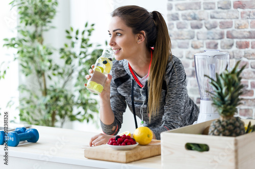 Fotomural Sporty young woman looking sideways while drinking lemon juice in the kitchen at home