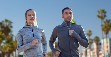 Wall Mural - fitness, sport and technology concept - happy couple running and listening to music in earphones over venice beach background in california