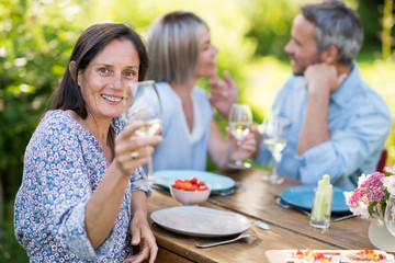 Wall Mural - In summer. a group of friends in their forties gathered around a table in the garden to share a meal. They toast with their glasses of wine to the camera.