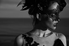 Black And White Dramatic Portrait Of Beautiful Young Woman In Dress With Black Flowers And With Black Lace Mask On Face And Floral Crown On Head. Sea Background
