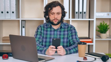 Fototapete - engineer sitting at the workspace using smartphone texting message and drinking coffee. handsome bearded man wearing casual shirt messaging in office