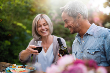  group of friends in their forties gathered around a table in the garden to share a bbq meal. A man offers red wine to a female friend