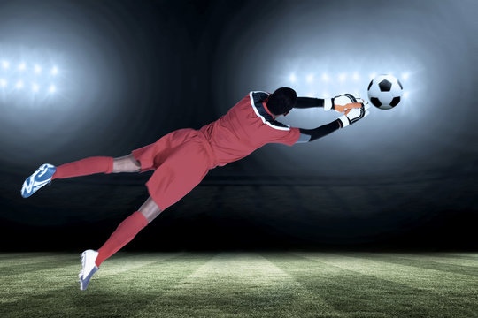 Goalkeeper in red making a save against football pitch under spotlights