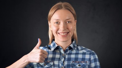 Poster - portrait young caucasian woman with blond hair posing on black background. attractive girl wearing casual bright shirt looking at the camera with friendly smile showing sign thumbs up