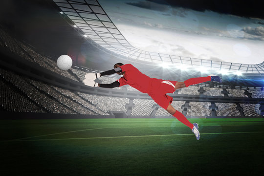 Goalkeeper in red making a save in a large football stadium with lights