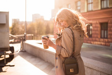 Walking Is A Spiritual Journey And A Reflection Of Living! Warm And Sunny Professional Photo Of The Young Smart Model Smiling And Drinking Coffee On The Street, While Smoothing Her Wavy Hair.