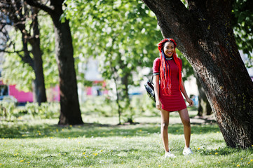 Wall Mural - Cute and slim african american girl in red dress with dreadlocks posed outdoor in spring park. Stylish black model.