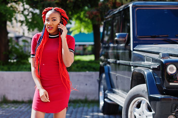 Wall Mural - Cute and slim african american girl in red dress with dreadlocks background black mafia suv car and speking on mobile phone. Stylish black model.