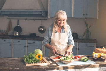 Enjoying household. Cheerful old woman is chopping cucumber and tomato for salad. She is listening to music from earphones and smiling