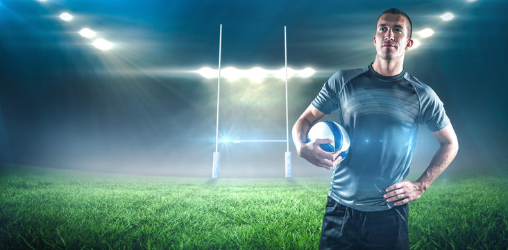 Rugby player holding ball with hand on hip against rugby stadium