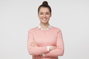 Horizontal portrait of young good-looking European Caucasian girl isolated on gray background standing with crossed arms, wearing eyeglasses and smiling happily at camera looking relaxed and calm