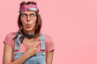 Indoor photo of amazed beautiful stylish hippie woman with headband and denim overalls, indicates at blank copy space for your advertising content, isolated over pink background. Wow, that`s amazing!