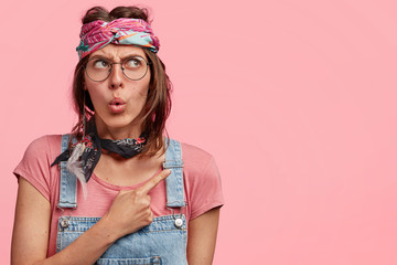 Wall Mural - Indoor photo of amazed beautiful stylish hippie woman with headband and denim overalls, indicates at blank copy space for your advertising content, isolated over pink background. Wow, that`s amazing!