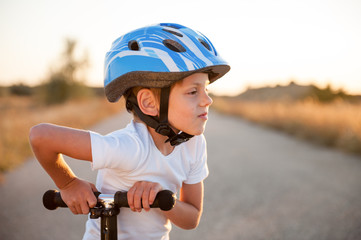 portrait or cute small child in sport helmet standing on abandoned road in Texas with scooter in warm summer sunset