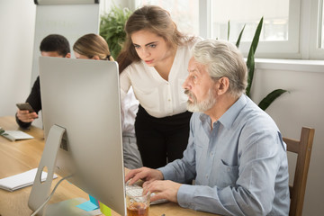 Female executive teaching senior colleague explaining computer work, corporate teacher helping focused aged manager with online task, young woman boss giving instructions to old employee at workplace