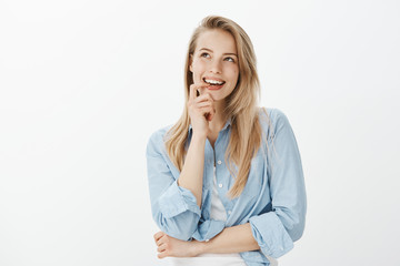 Wall Mural - Creative good-looking female designer with blond hair, biting finger, looking up and smiling curiously while having great idea, standing dreamy over gray background, having interesting plan